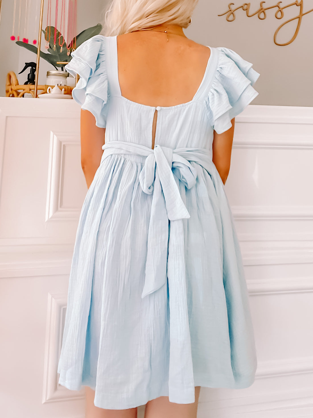 Light blue mini dress with ruffled sleeves and a tie waist, shown from the back against a white wall.
