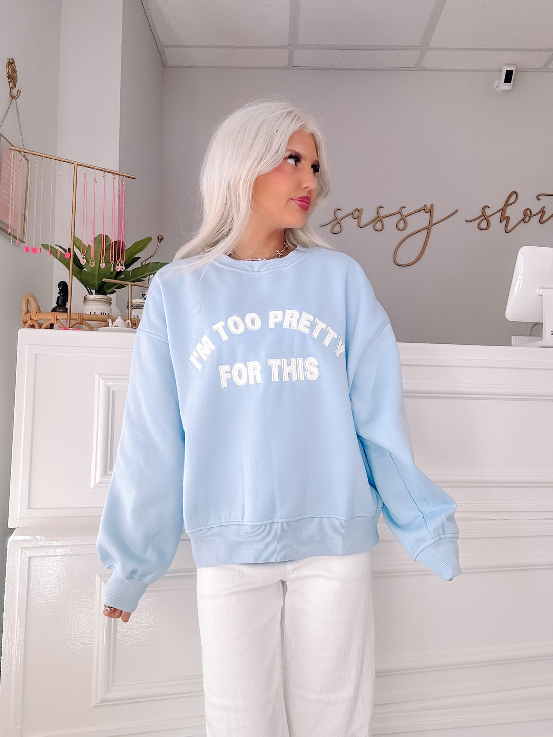 Light blue crewneck sweatshirt with "I'm too pretty for this" text, modeled in a shop.
