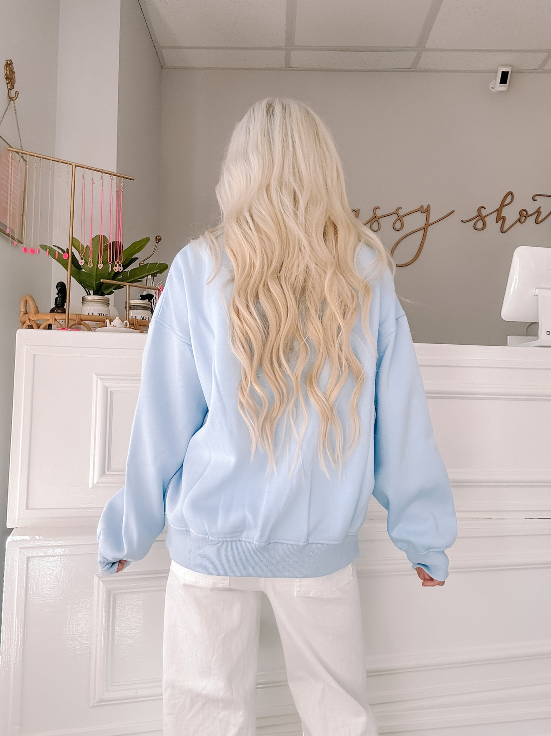 Light blue crewneck sweatshirt on a model, shown from behind in a shop setting.

