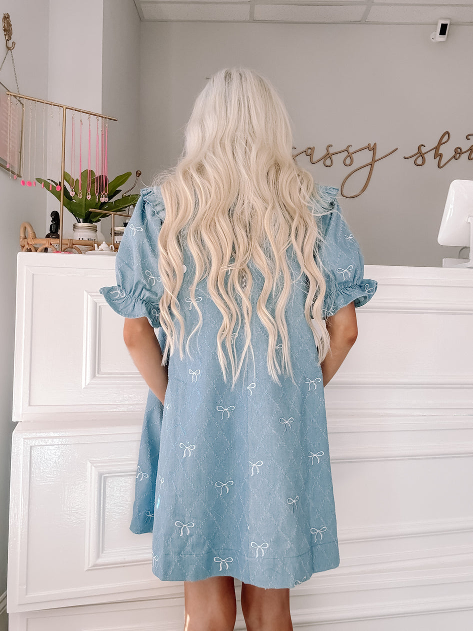 Light blue denim dress with embroidered white bows, puff sleeves, and a relaxed fit.
