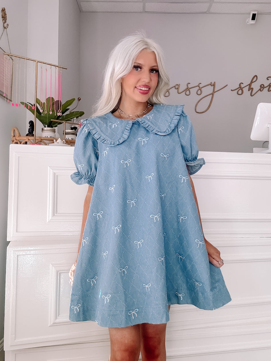 Light blue denim mini dress with embroidered white bows, peter pan collar, and short puff sleeves.
