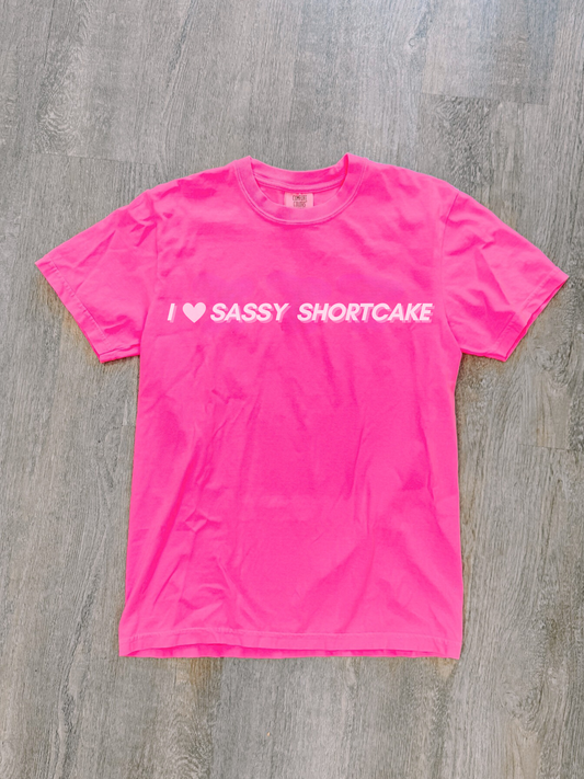 Flatlay of hot pink tee with white "I heart Sassy Shortcake" graphic, laid neatly on light floor.