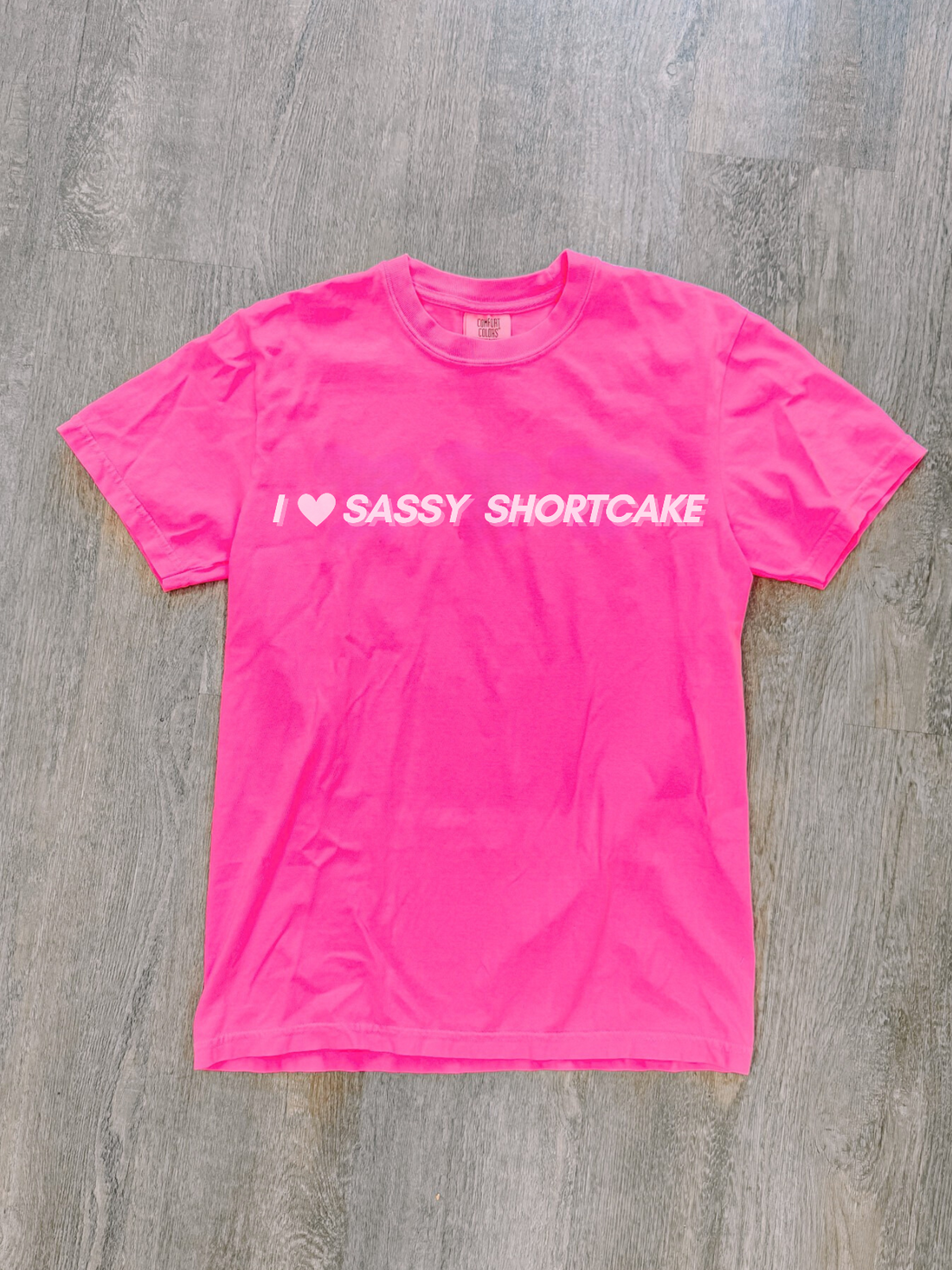 Flatlay of hot pink tee with white "I heart Sassy Shortcake" graphic, laid neatly on light floor.