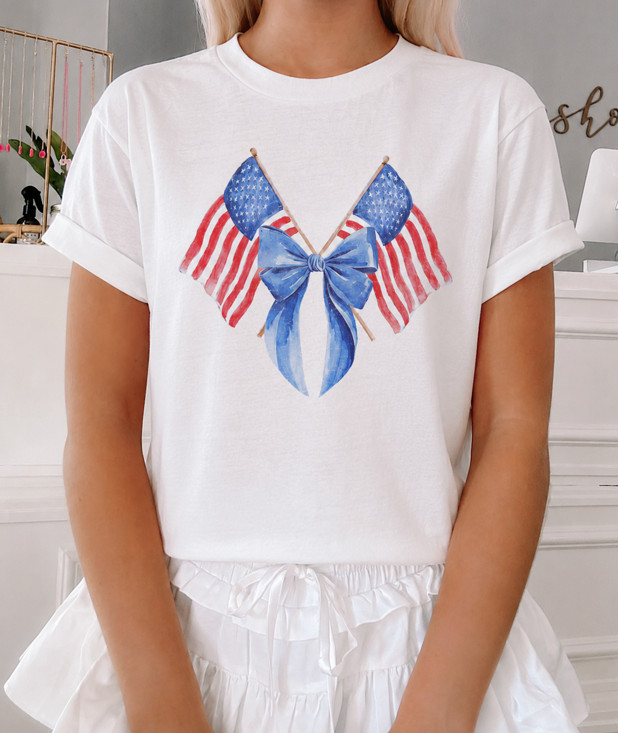 Model wearing The Liberty Ribbon Tee from Sassy Shortcake, a white cotton t-shirt featuring a watercolor graphic of crossed American flags tied with a blue bow, paired with a white ruffled skirt.