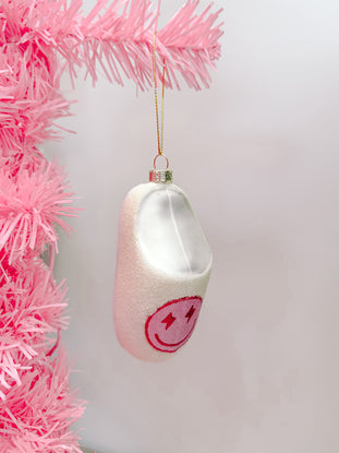 White slipper Christmas ornament with pink smiley face, hanging on a pink tree.
