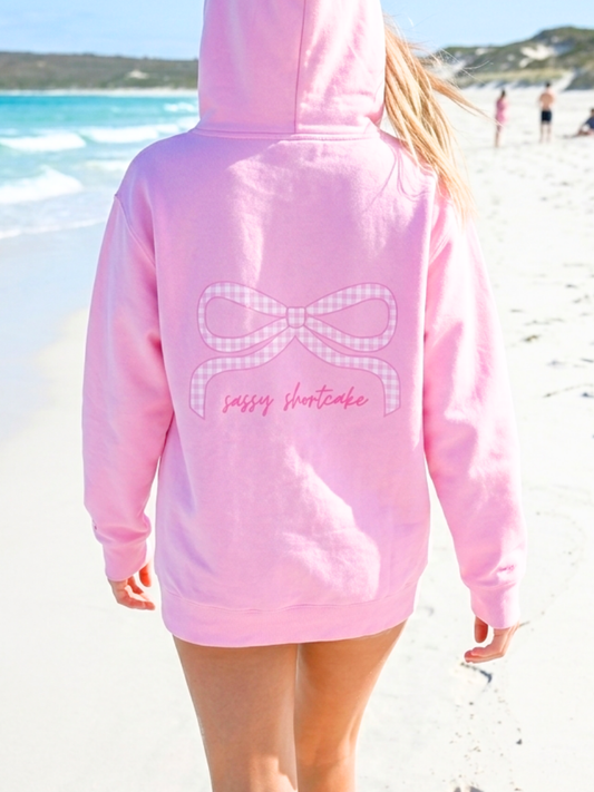 Sassy Gingham Bow Hoodie from Sassy Shortcake - Person wearing a pink hoodie with a bow design and text on a beach.
