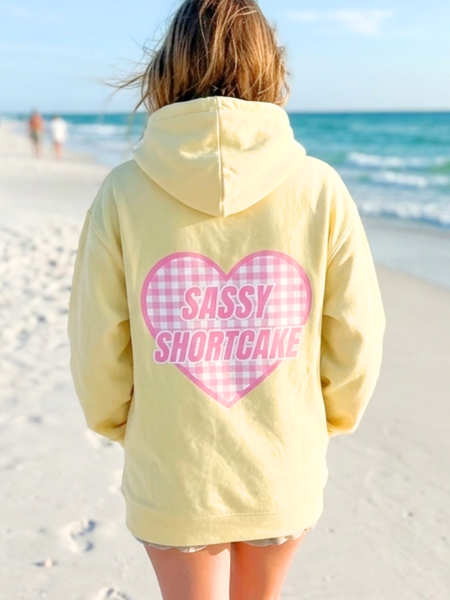 Sassy Sun Love Hoodie from Sassy Shortcake - Person wearing a yellow hoodie with 'Sassy Shortcake' on a beach