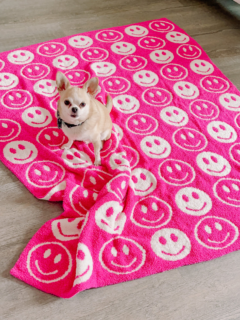 Pink fluffy blanket with white smiley faces, a small dog sitting on it.
