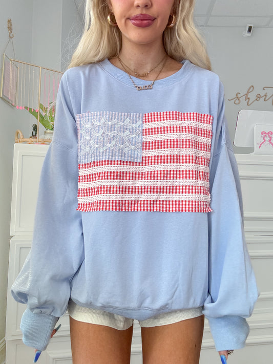 Model wearing blue oversized crewneck with American flag patchwork and shiny bow details.