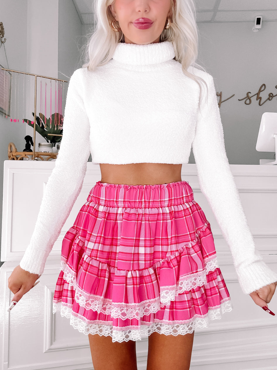 Pink plaid mini skirt with white lace trim, layered ruffles, and a fuzzy white sweater.
