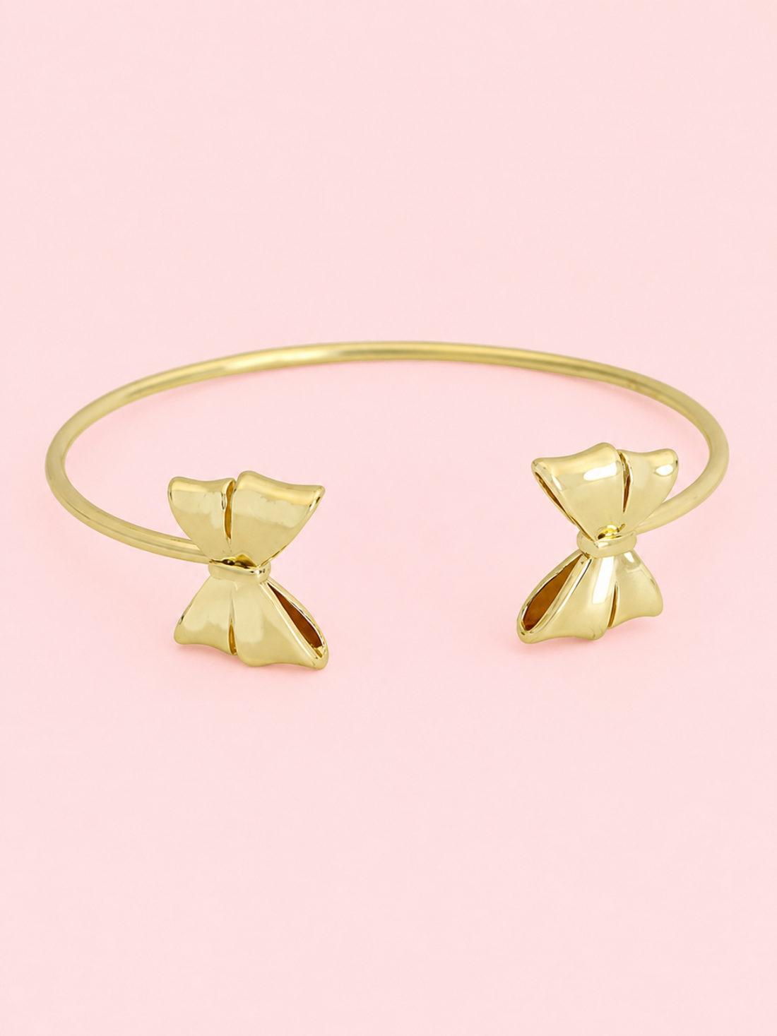 Wrapped In Gold Bow Cuff from Sassy Shortcake: Gold open cuff bracelet with bow details on each end displayed on a light pink background.
