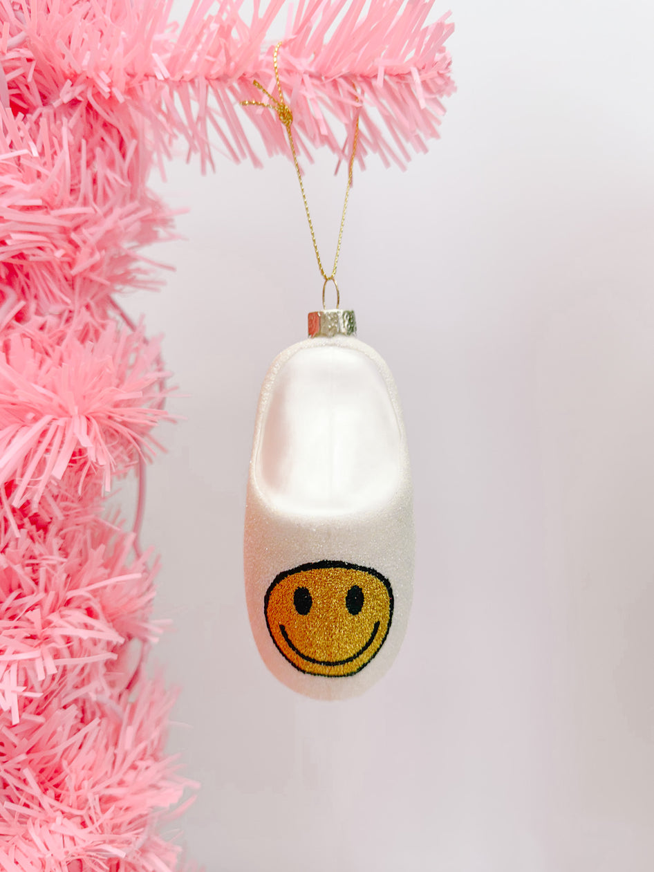 White slipper Christmas ornament with a gold smiley face, hanging on a pink tree.
