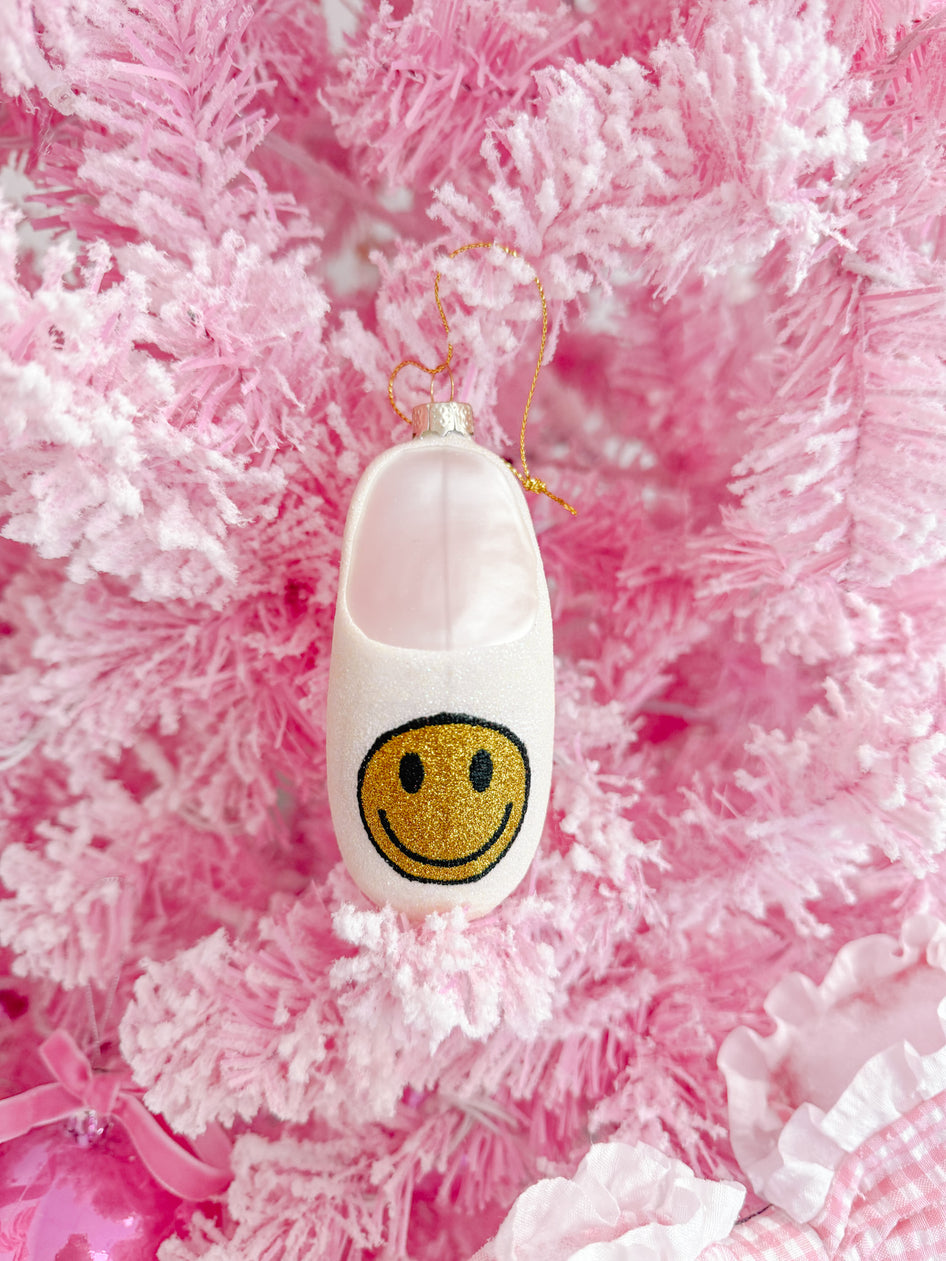 White glitter slipper Christmas ornament with gold smiley face, hanging on a pink tree.
