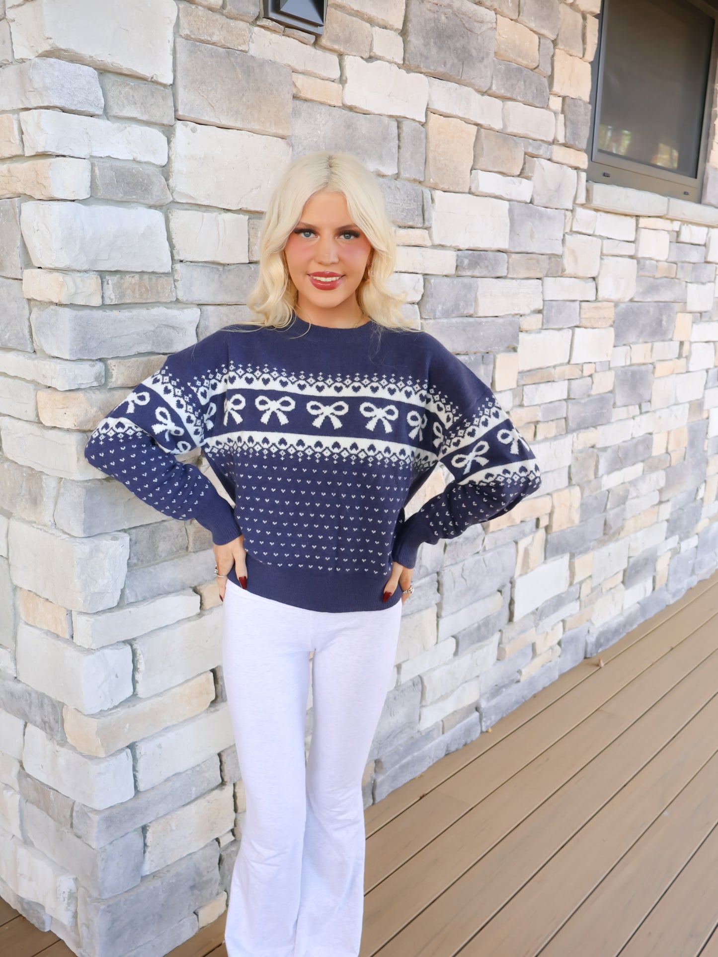 Woman modeling the Alpine Angel Navy Bow Sweater, a navy knit with a white bow and heart pattern for seasonal charm.