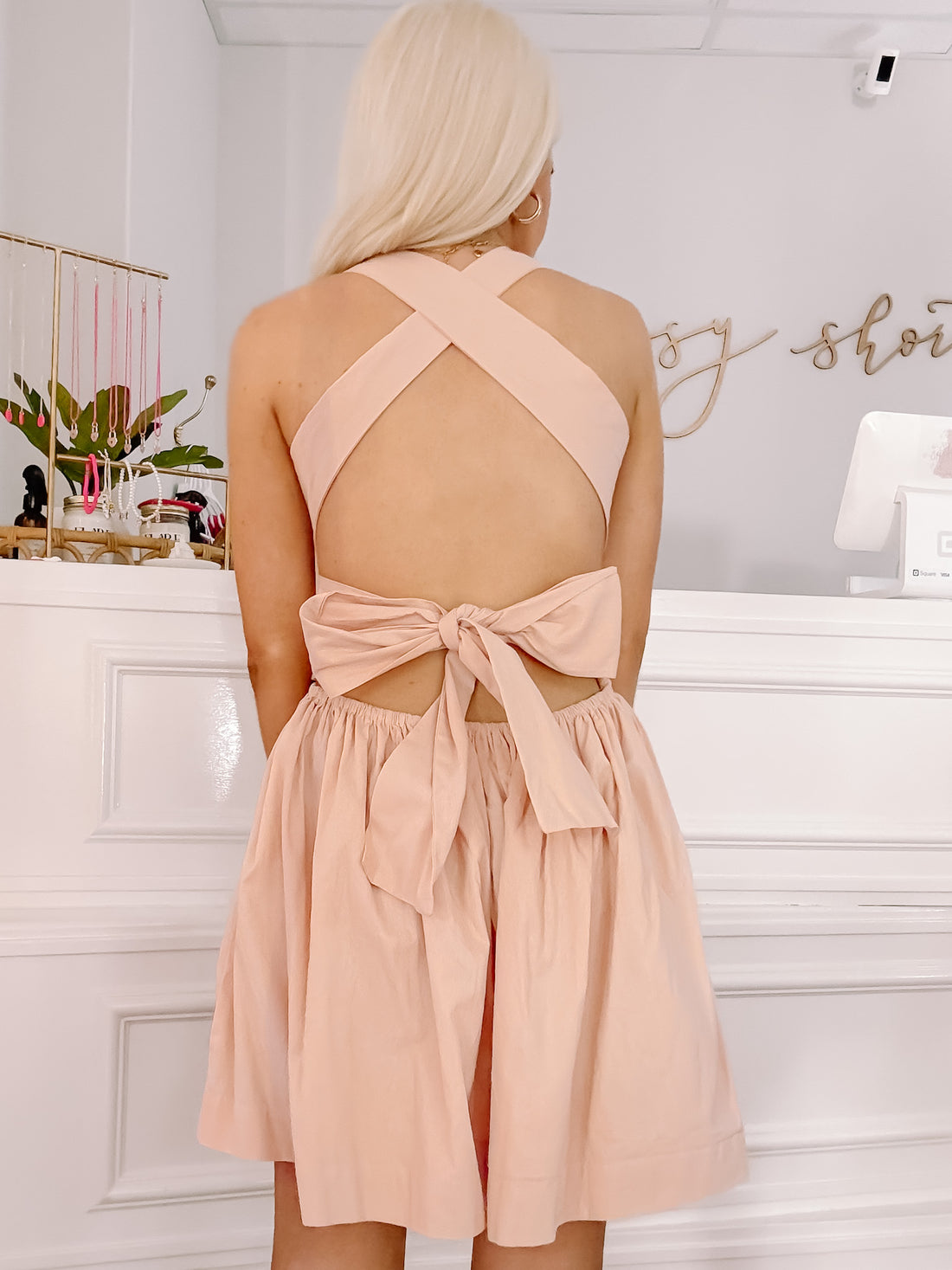 Blush pink mini dress with crisscross straps and a bow tie back detail, shown in a boutique setting.
