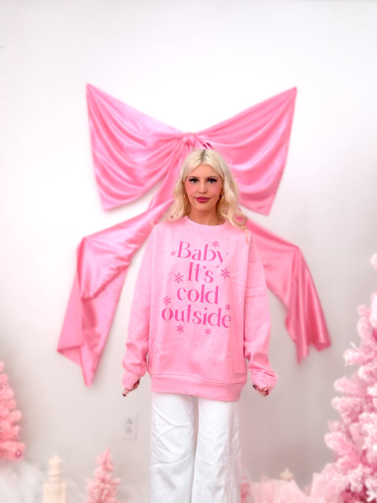 Model wearing a pink crewneck sweatshirt with the text "Baby It's Cold Outside" and snowflakes in hot pink, showing the fleece lining detail.