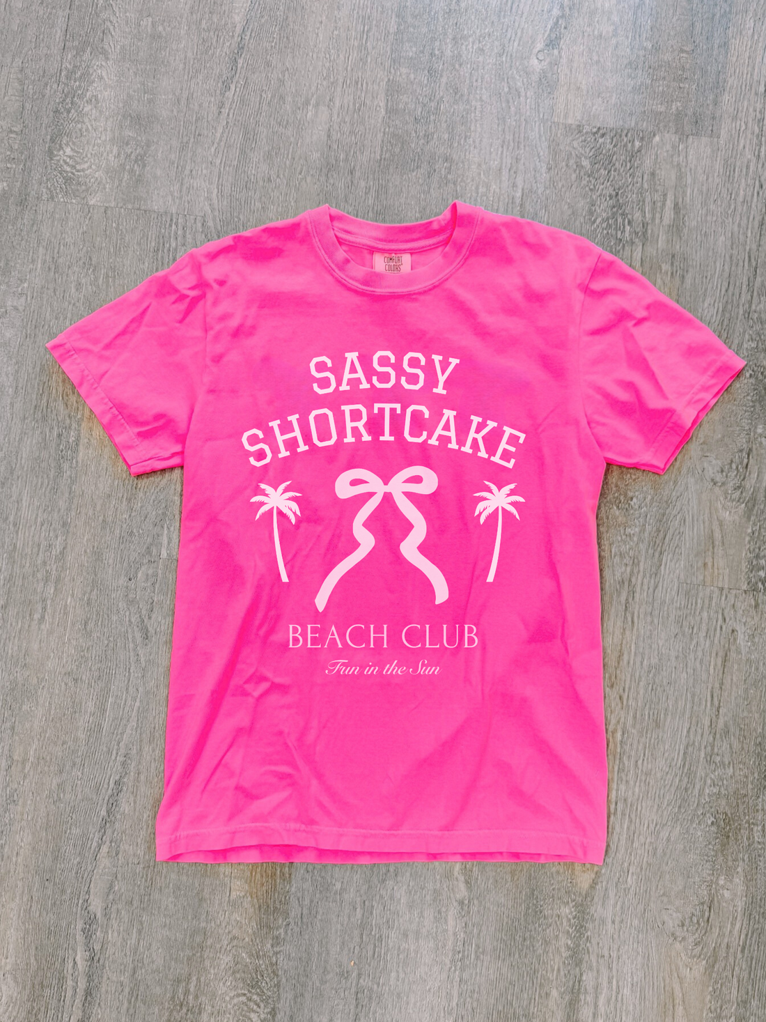 Pink Sassy Shortcake Beach Club Merch bow tee neatly laid flat on whitewashed wood floor.