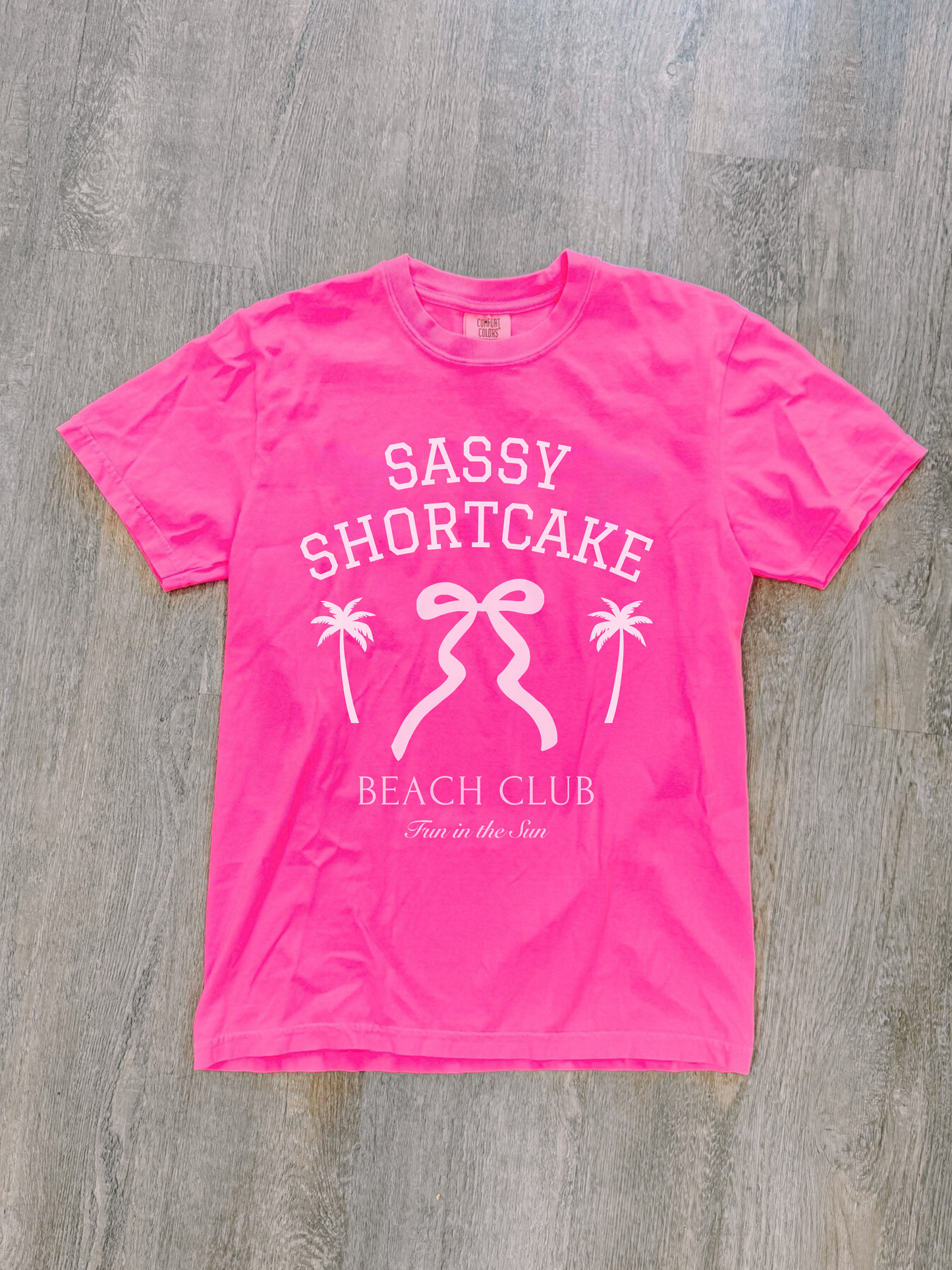 Pink Sassy Shortcake bow tee neatly laid flat on whitewashed wood floor.