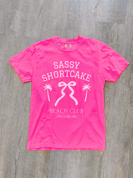 Pink Sassy Shortcake bow tee neatly laid flat on whitewashed wood floor.
