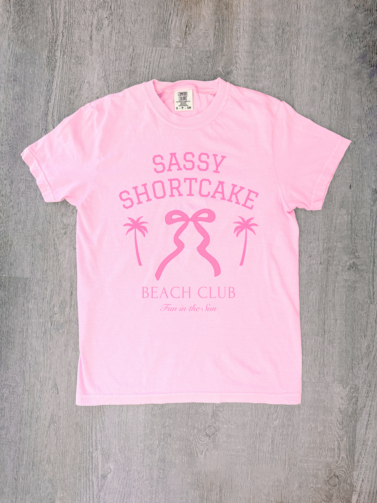 Flatlay of soft pink tee featuring bow logo, shot from above.
