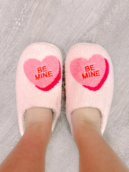 Pink fuzzy slippers with "Be Mine" heart design, worn on a wood floor.
