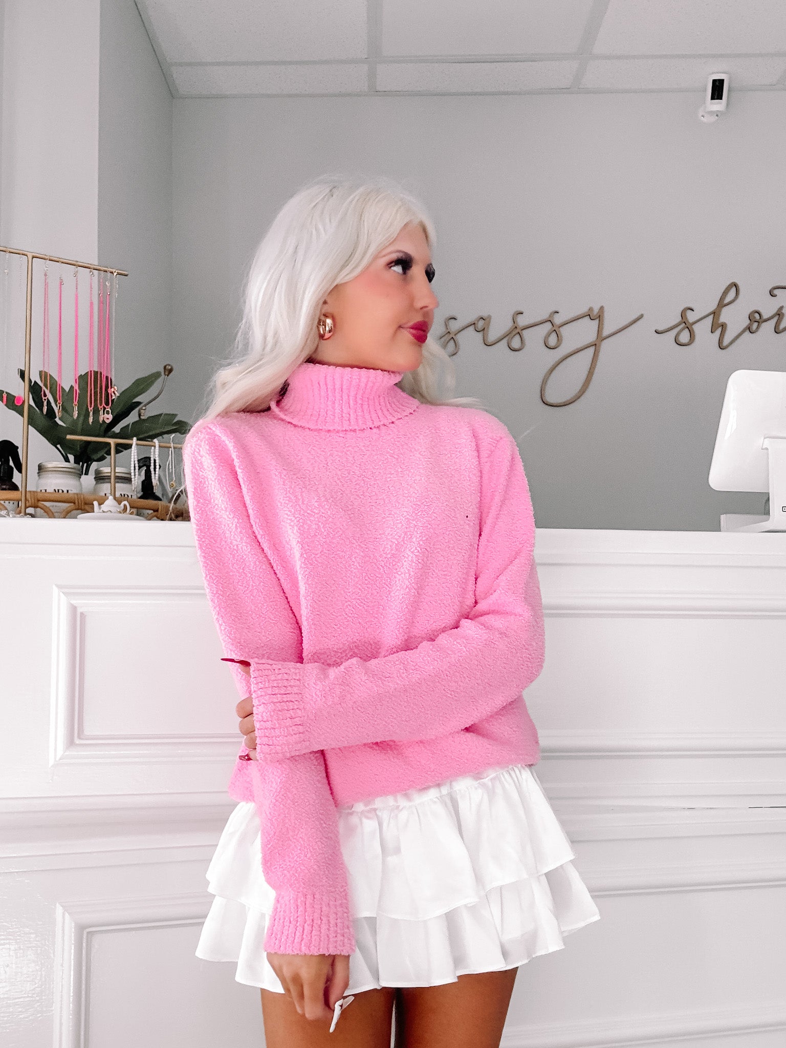 Pink fluffy turtleneck sweater styled with a white tiered ruffle skirt in a boutique setting.
