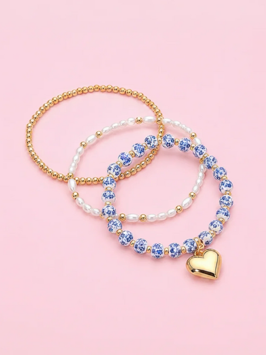 Three beaded stretch bracelets arranged in overlapping circles on a soft pink background. One bracelet is gold-tone beads, one is white faux pearls with gold beads, and one is blue-and-white floral beads with a dangling gold heart charm. Blue Belle bracelet stack from sassy shortcake