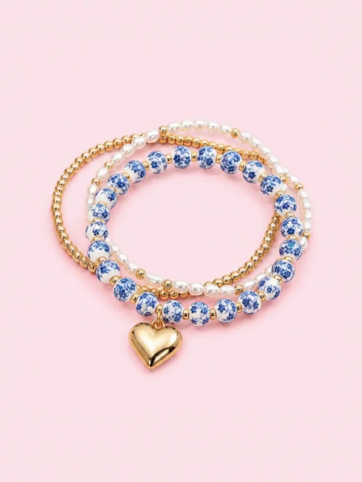 A set of three beaded stretch bracelets with a gold heart charm, featuring blue and white ceramic beads, white pearl-like beads, and gold beads, displayed on a solid pink background. Blue Belle Bracelet stack - sassy shortcake