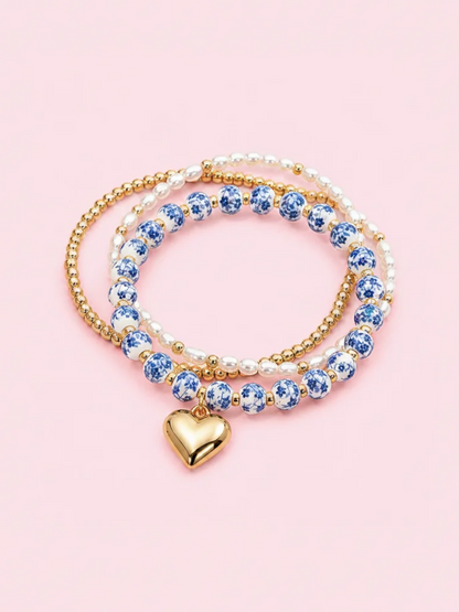 A set of three beaded stretch bracelets with a gold heart charm, featuring blue and white ceramic beads, white pearl-like beads, and gold beads, displayed on a solid pink background. Blue Belle Bracelet stack - sassy shortcake