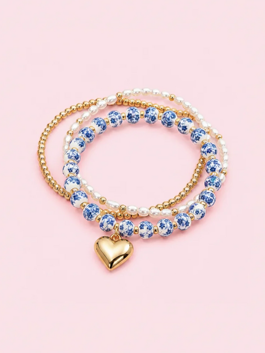 A set of three beaded stretch bracelets with a gold heart charm, featuring blue and white ceramic beads, white pearl-like beads, and gold beads, displayed on a solid pink background. Blue Belle Bracelet stack - sassy shortcake