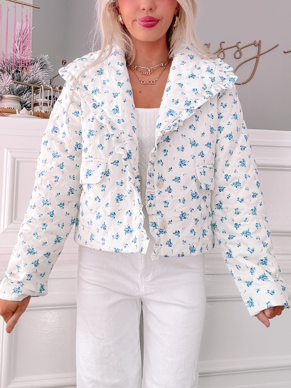 White quilted jacket with a delicate blue floral print, featuring ruffled detailing at the collar.
