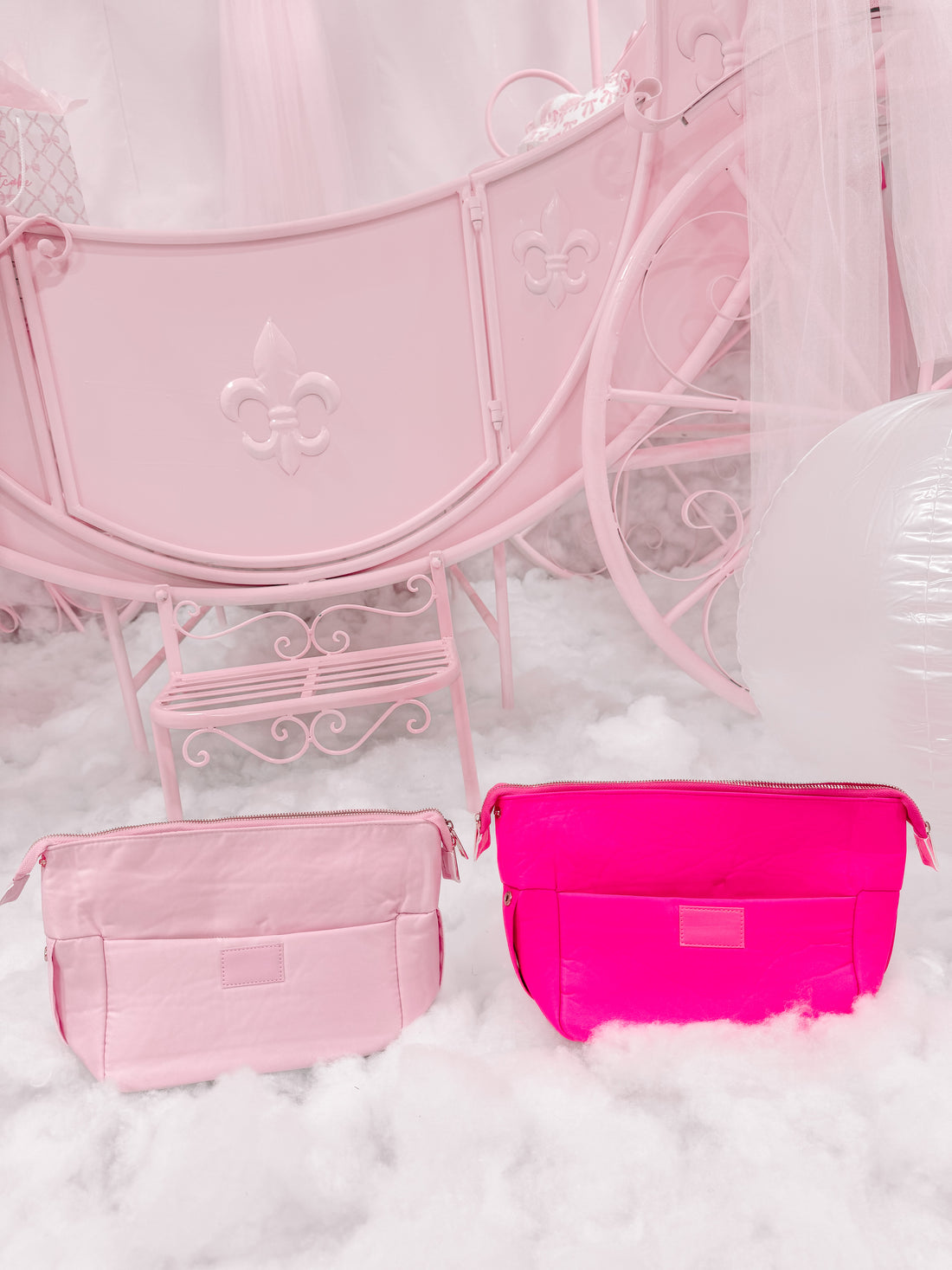 Pink and hot pink zippered pouches displayed on fluffy white surface near a pastel pink carriage.
