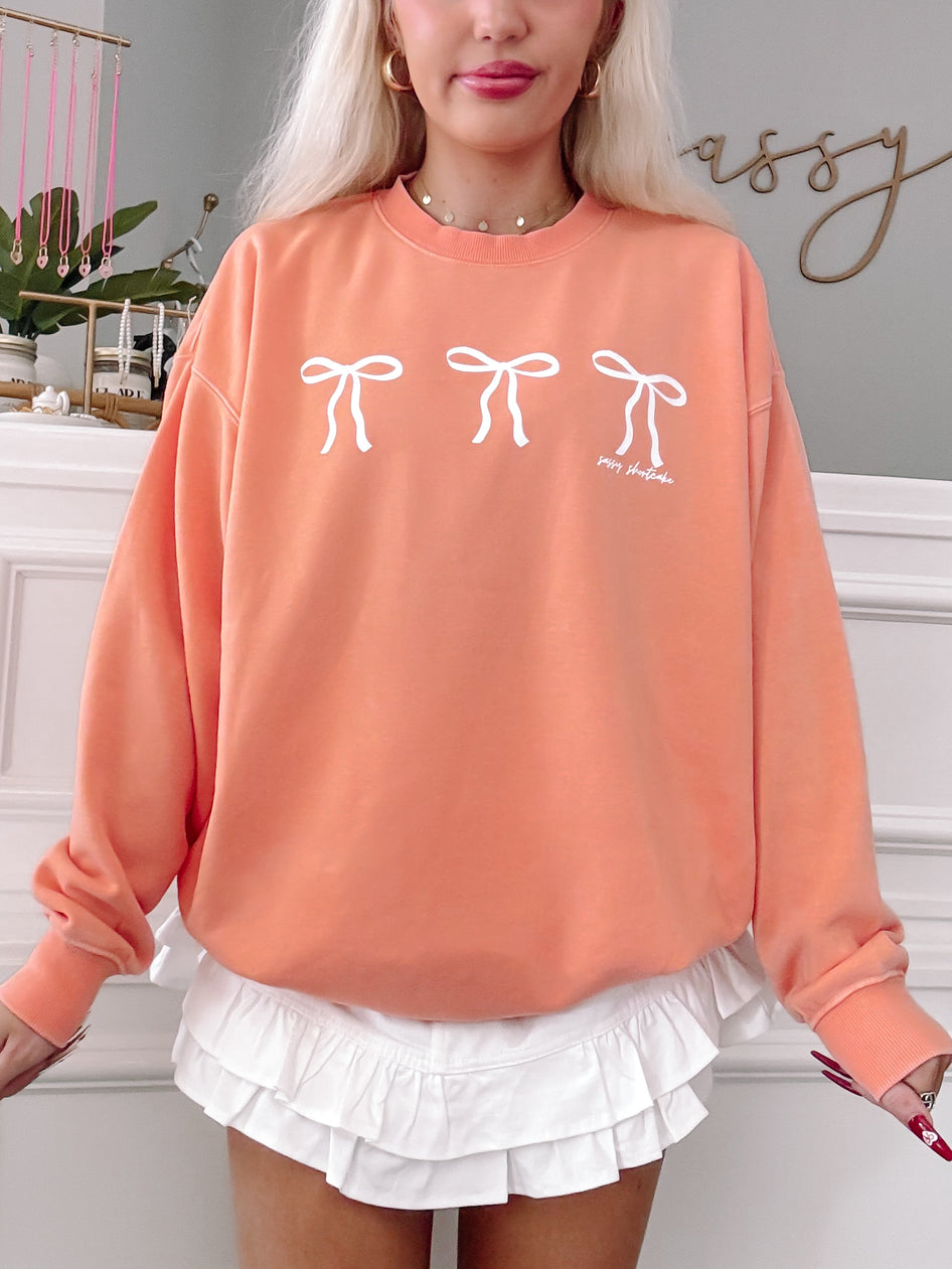 Peach crewneck sweatshirt with three white bows, styled with a white ruffled skirt.
