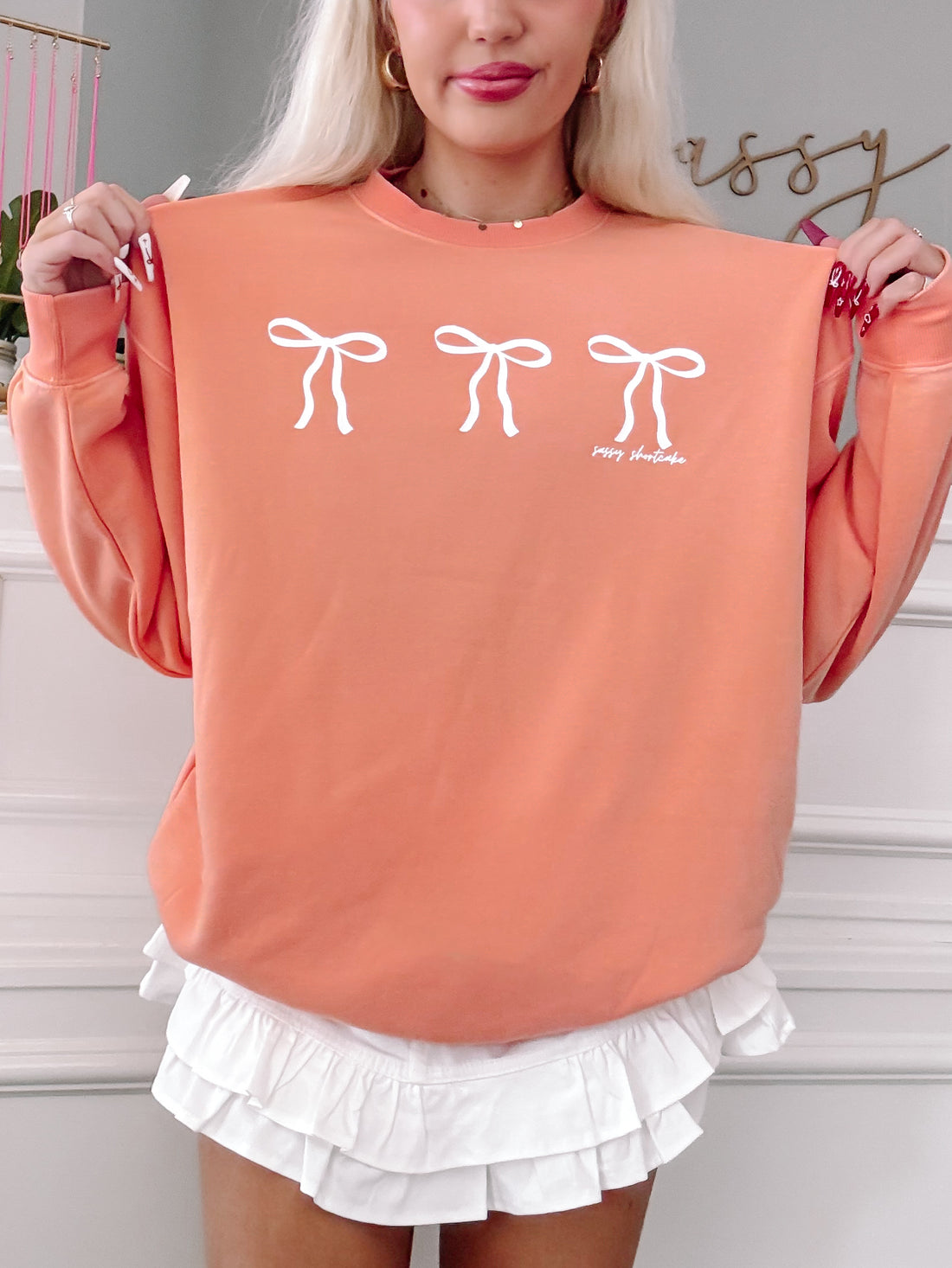 Peach crewneck sweatshirt featuring three white bows, styled with a white ruffled skirt.
