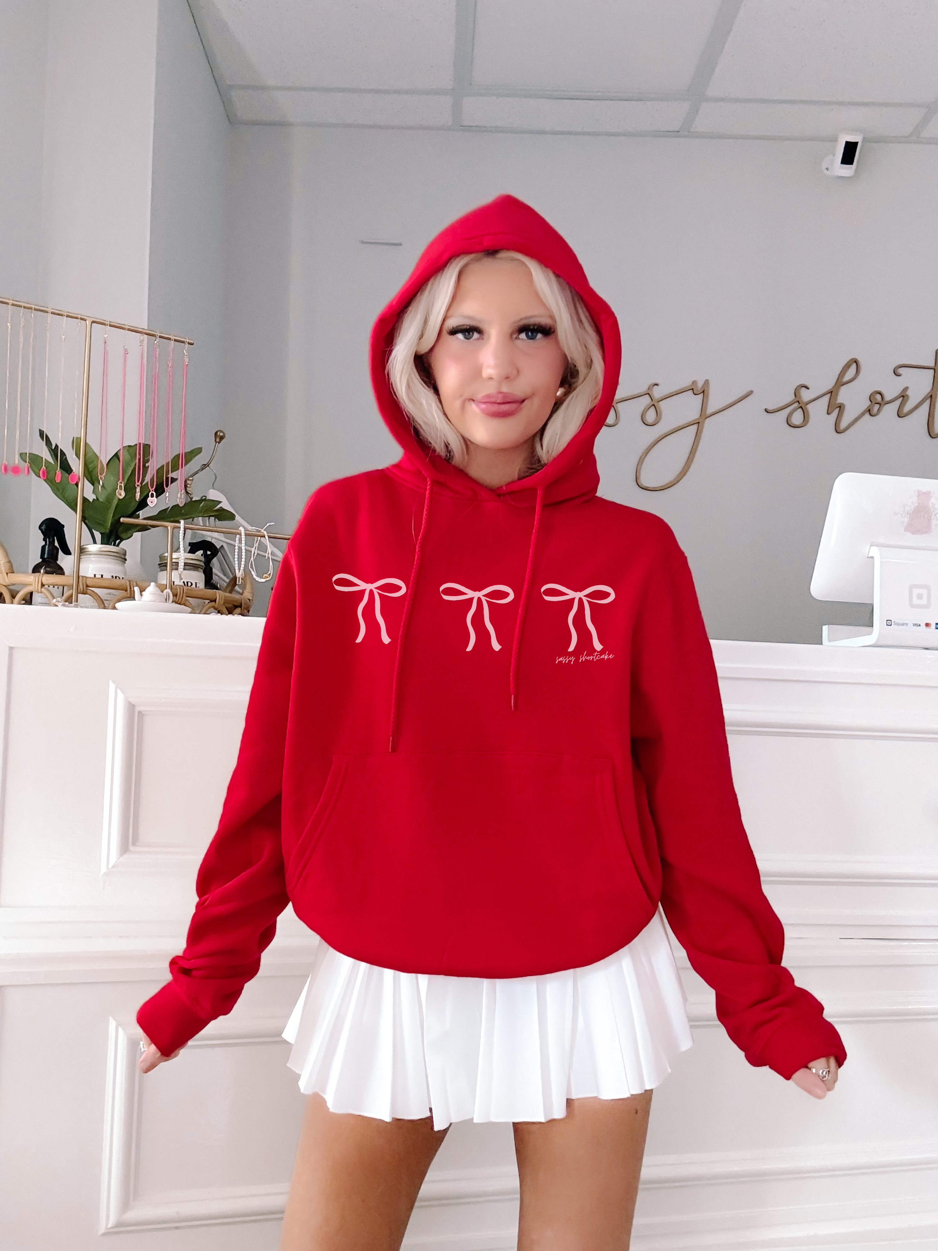 Red hoodie featuring delicate pink bows, styled with a white pleated skirt in a bright interior.
