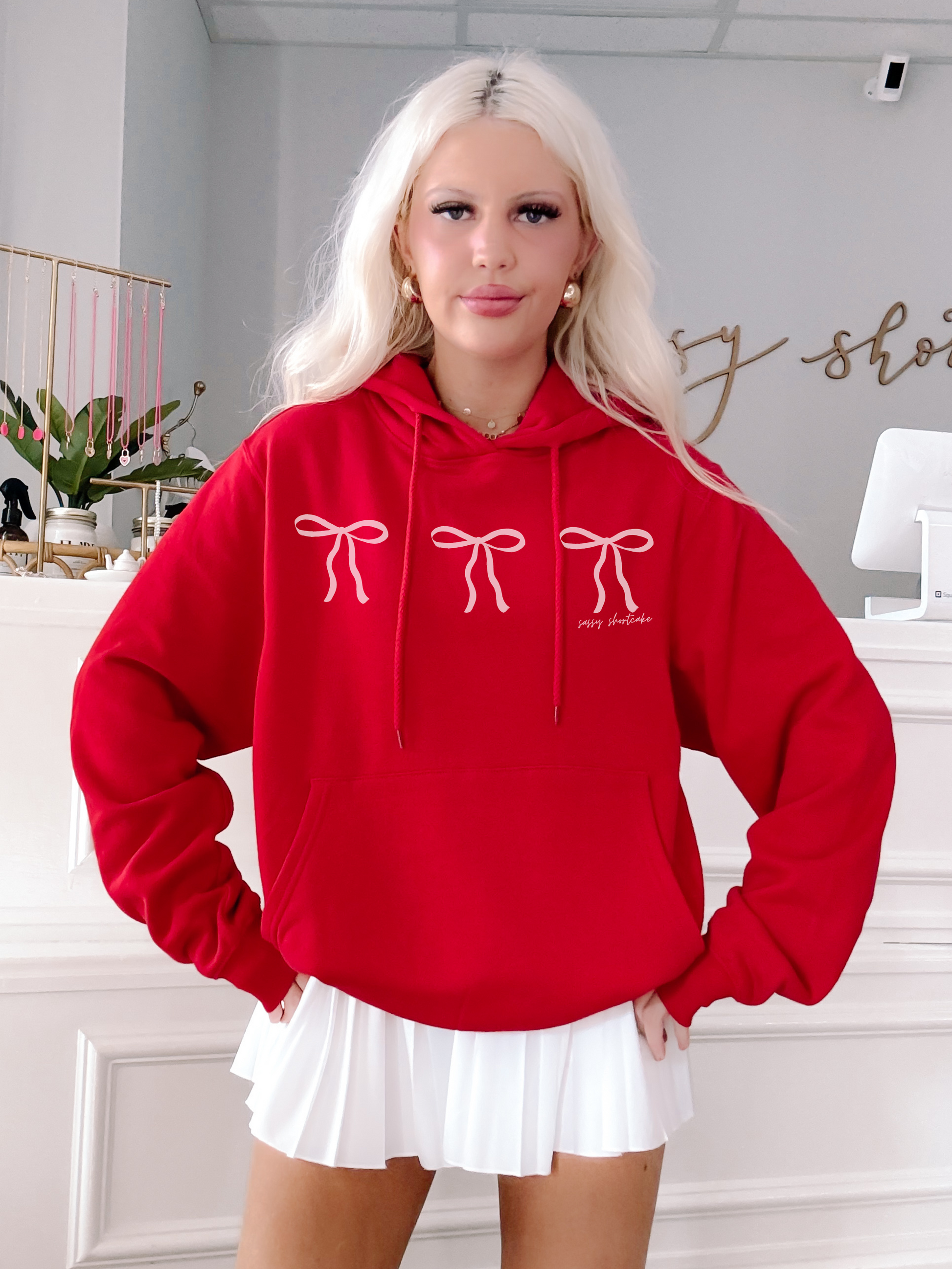 Red hooded sweatshirt featuring a trio of pink bows, styled with a white pleated skirt.
