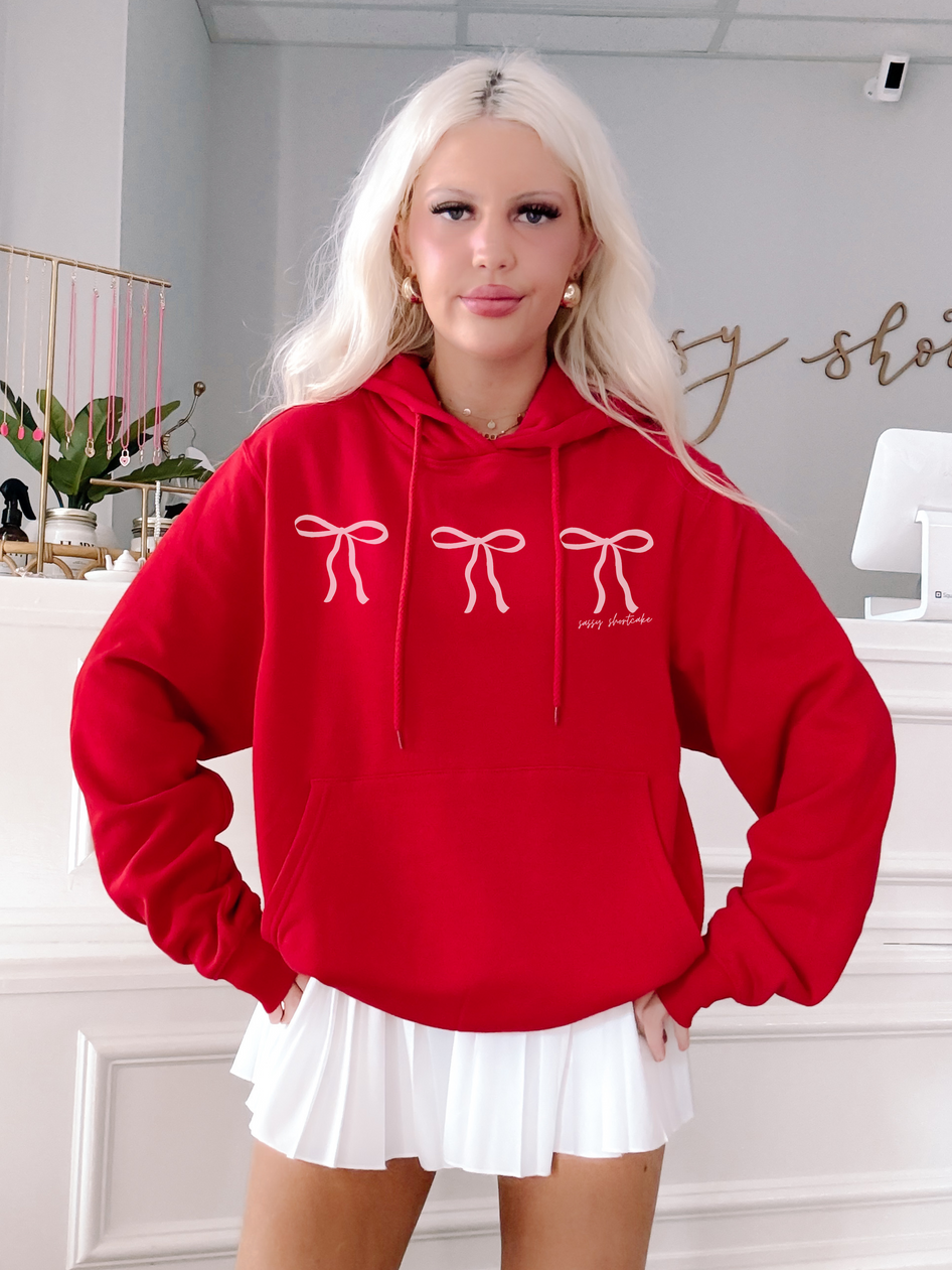 Red hoodie featuring a trio of pink bows, modeled against a light background.
