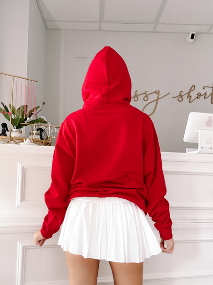 Red hoodie worn with a white pleated skirt, shown from behind in a bright retail setting.
