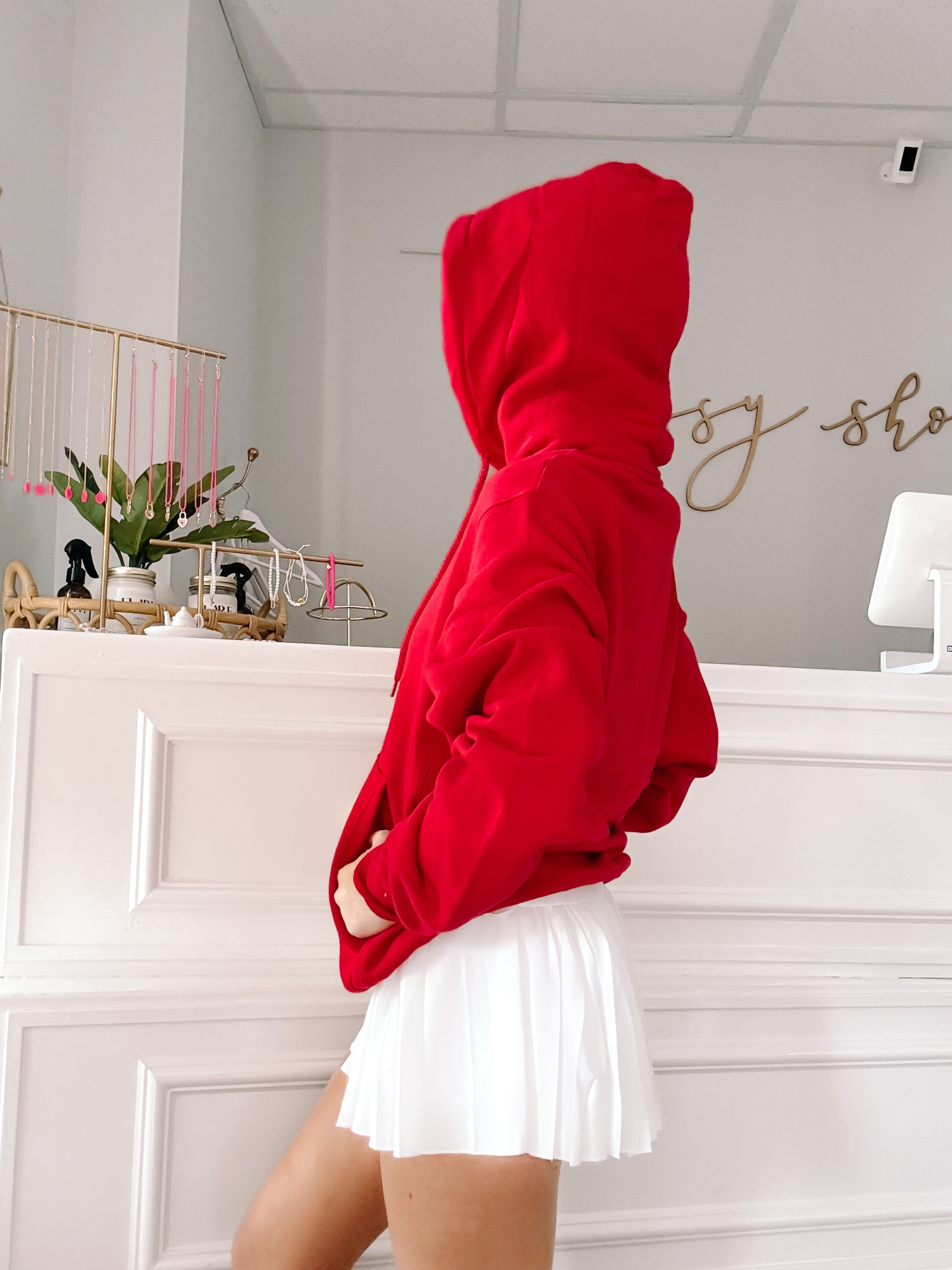 Red hooded sweatshirt styled with a white pleated skirt, shown in a boutique setting.
