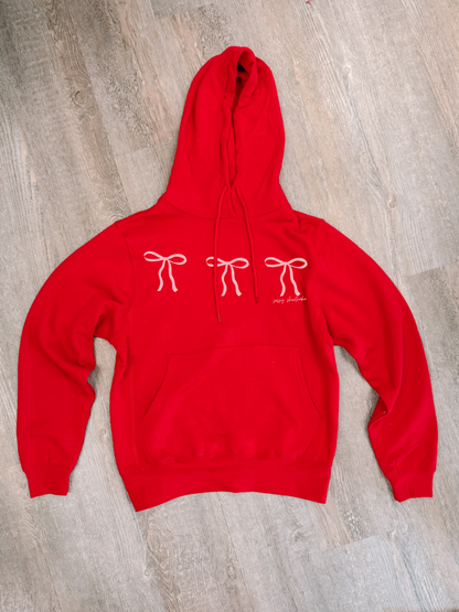 Red hooded sweatshirt with pink bow design, shown laid flat on wood.
