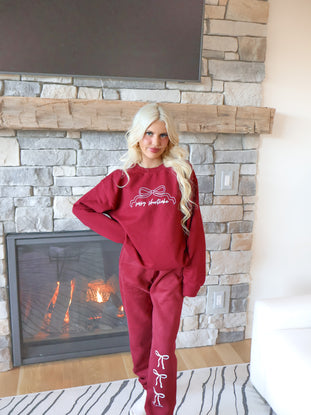 Model wearing burgundy sweatpants with an elastic waistband, pockets, and a soft micro-fleece lining, showing the three white bow design.