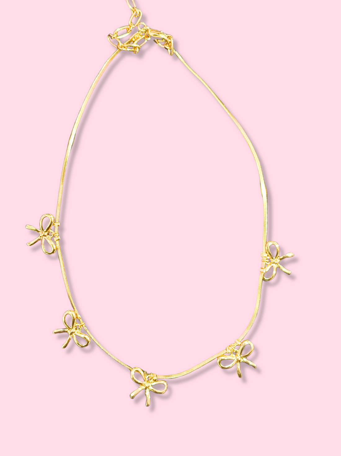 Gold necklace with delicate bow charms on a pink background.
