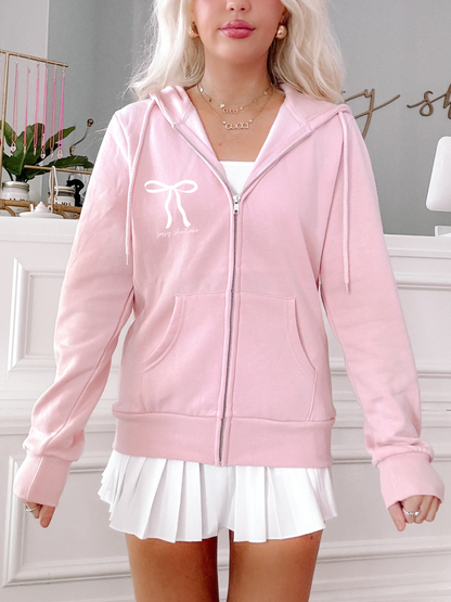 Pink zip-up hoodie featuring a bow and text detail, modeled against a light background.
