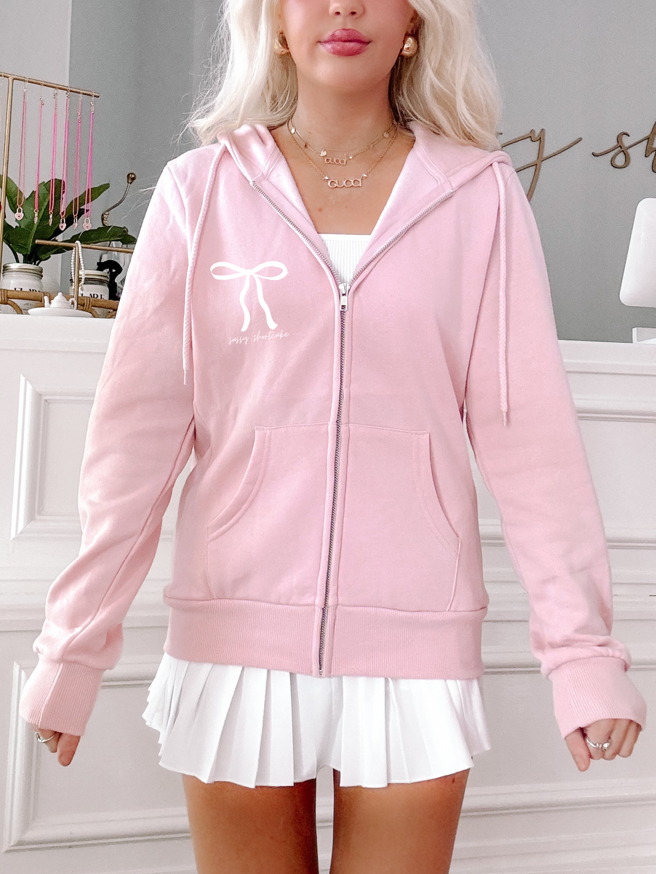 Baby pink zip-up hoodie featuring a bow and text detail;  model wearing with white pleated skirt.
