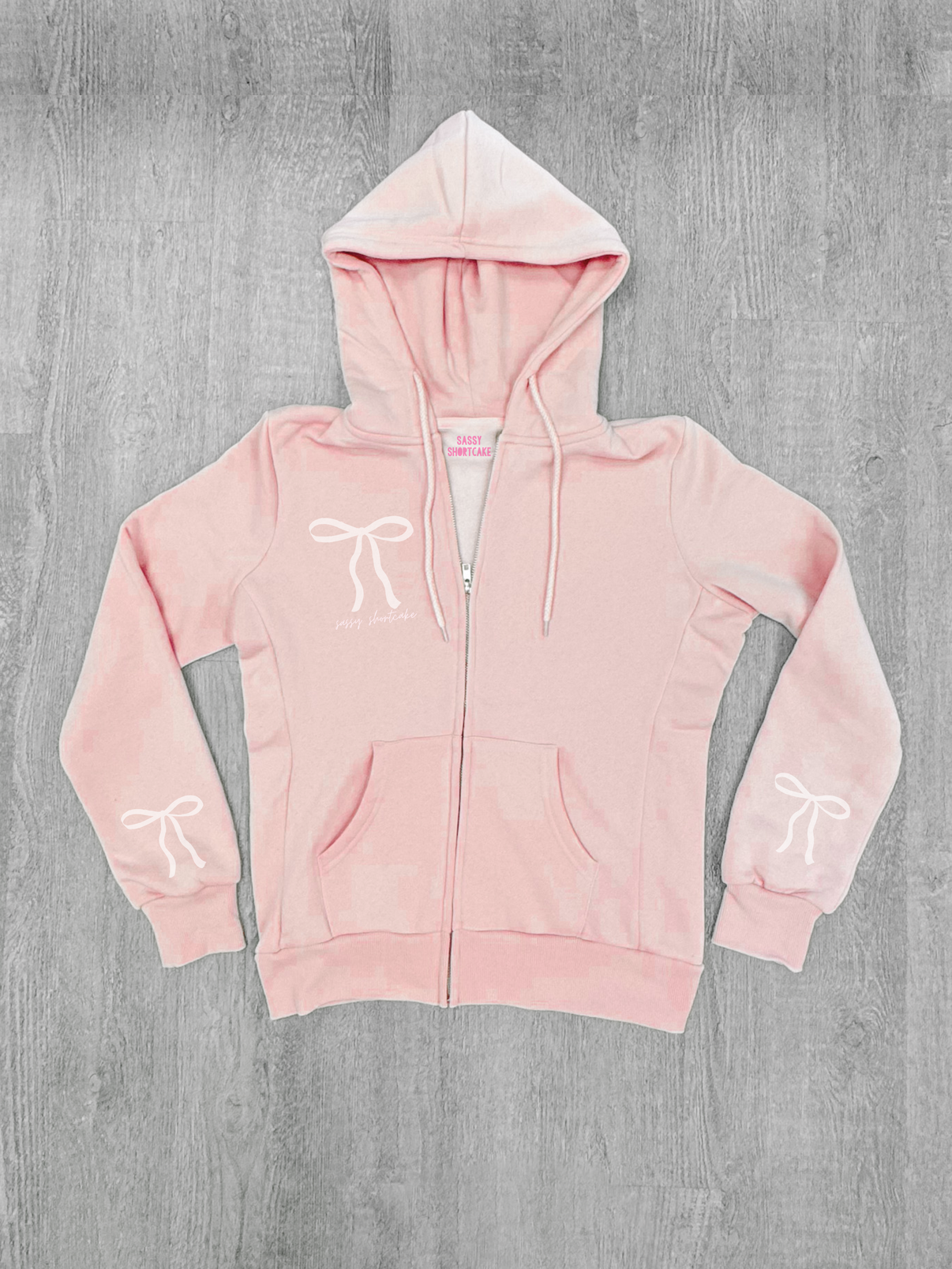 Baby pink zip-up hoodie with bow details, shown laid flat on a gray wood background.
