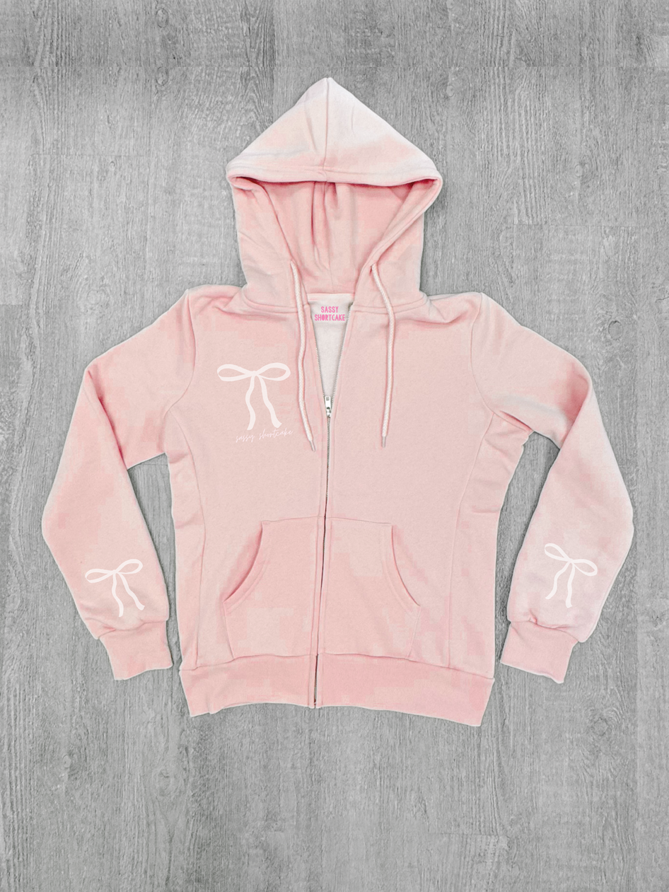 Baby pink zip-up hoodie with bow details, shown on a gray wood background.
