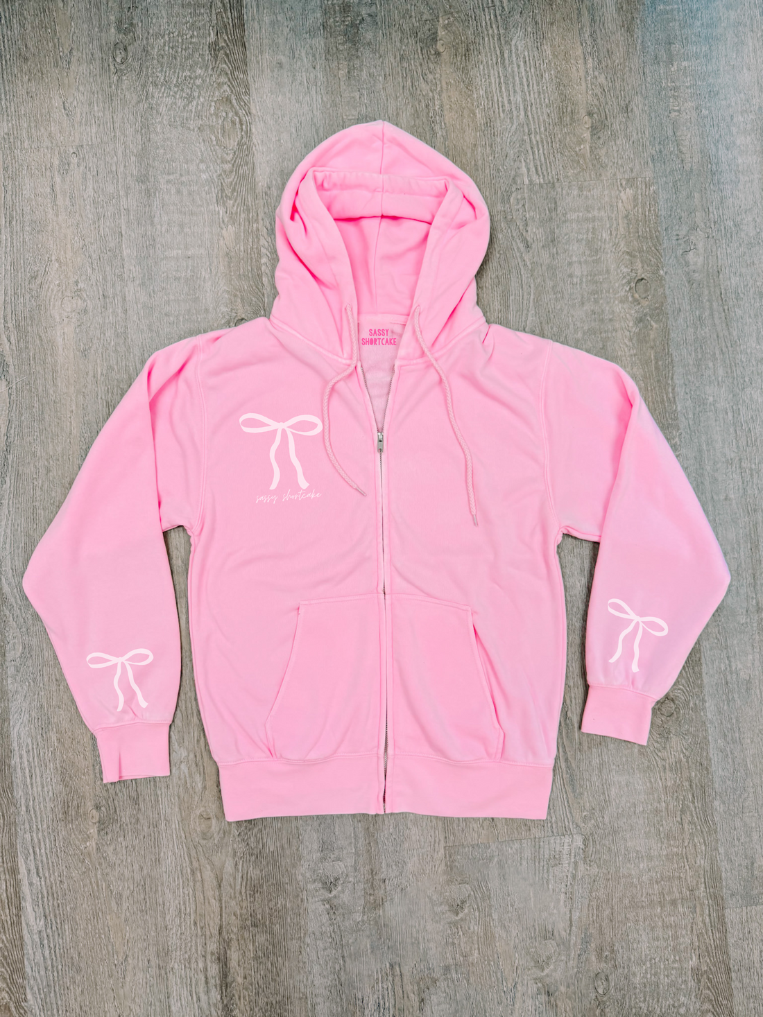 Bubblegum pink zip-up hoodie with bow details, shown flat lay on wood.
