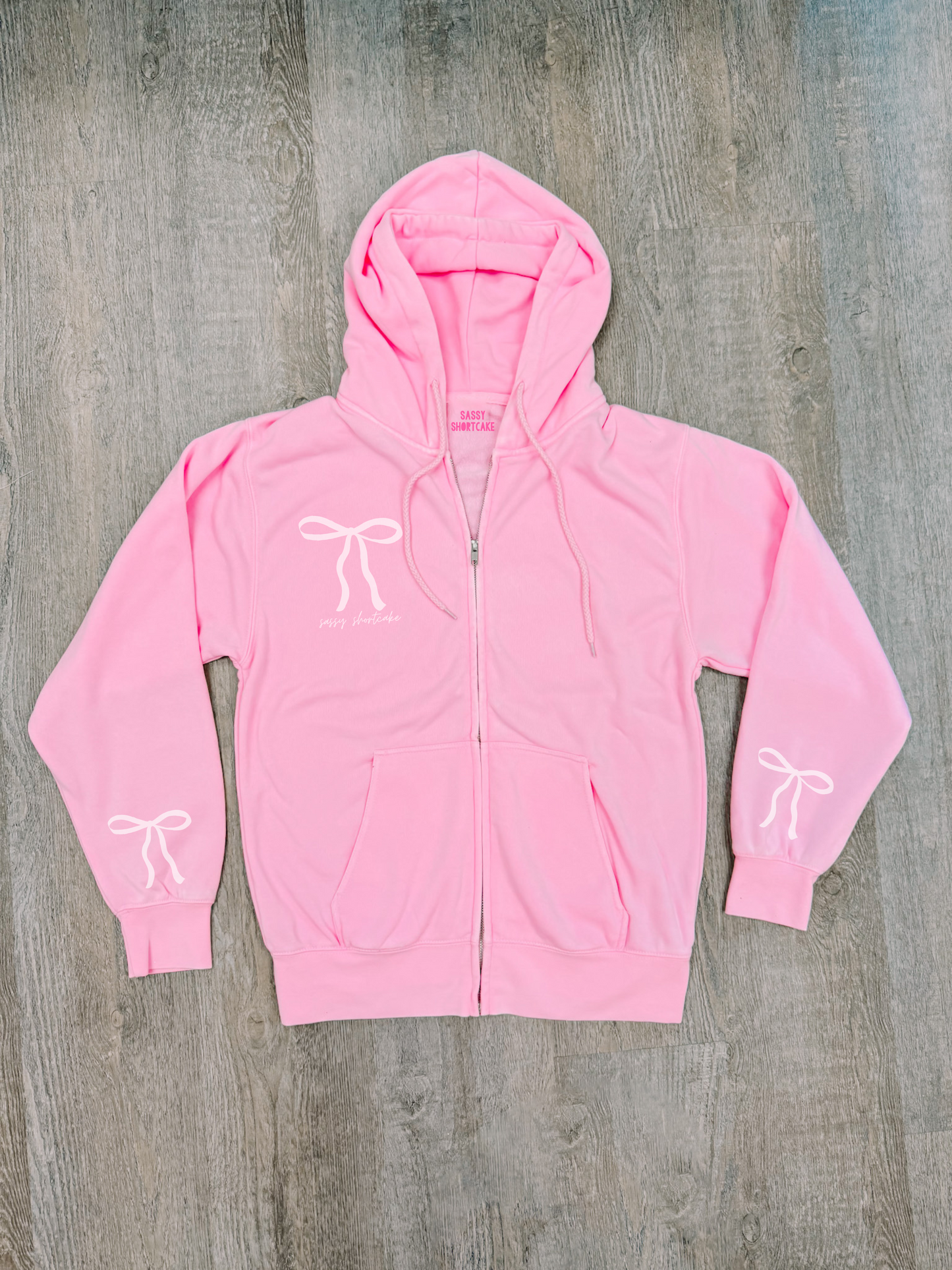 Bubblegum pink zip-up hoodie with bow details, shown flat lay on wood.
