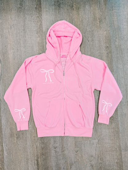 Bubblegum pink zip-up hoodie with bow details, shown flat lay on wood.
