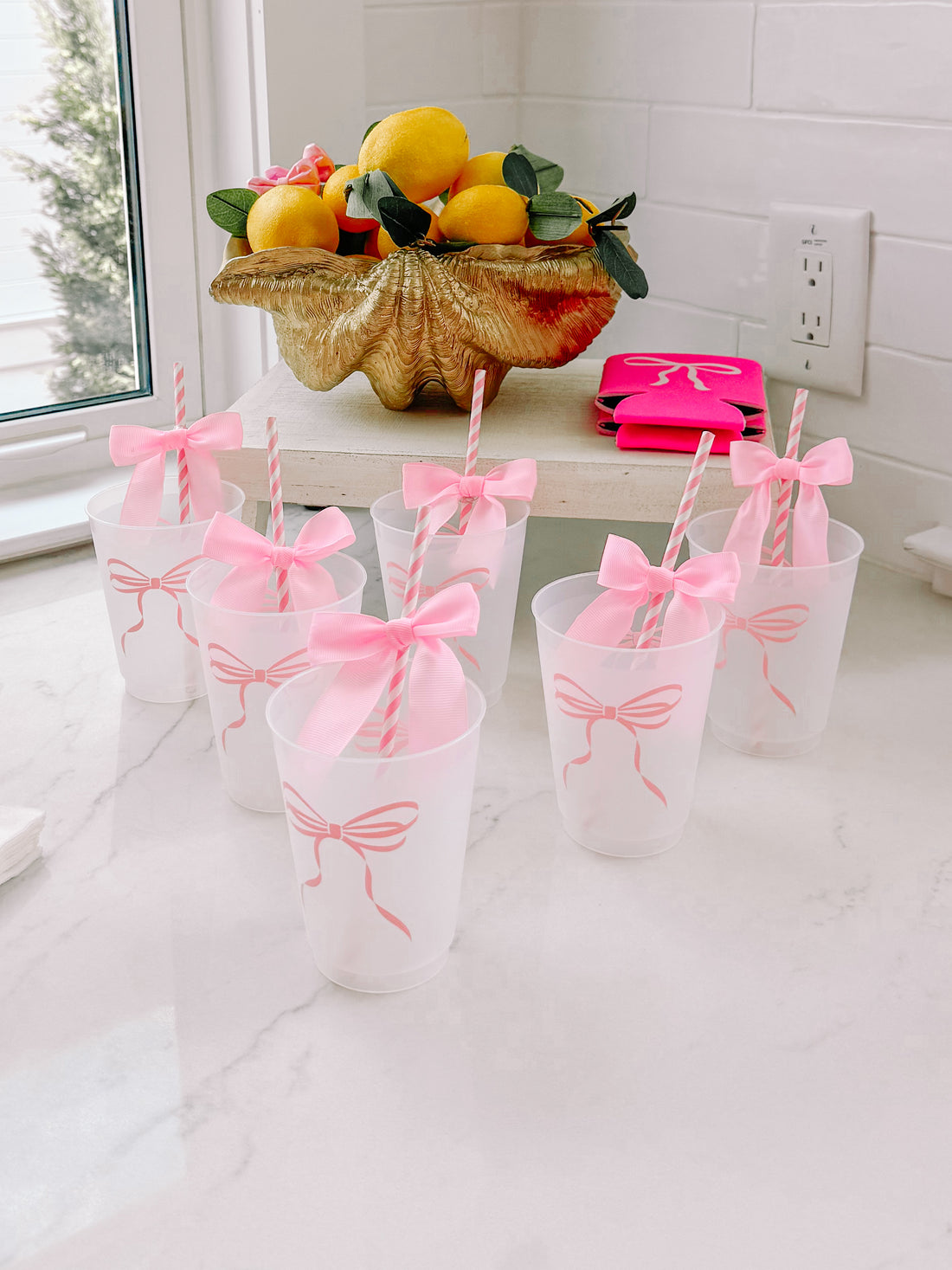 Frosted plastic cups decorated with pink bows, paired with striped straws, arranged on a marble surface.
